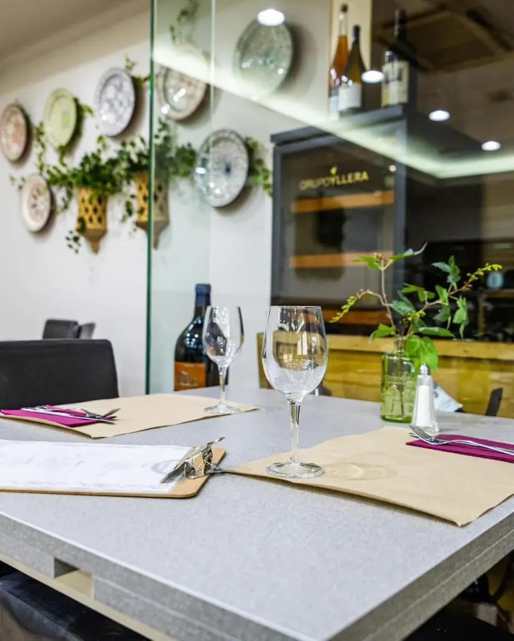Mesa de restaurante con copas, botella de vino y cubiertos; platos decorativos y plantas en la pared.