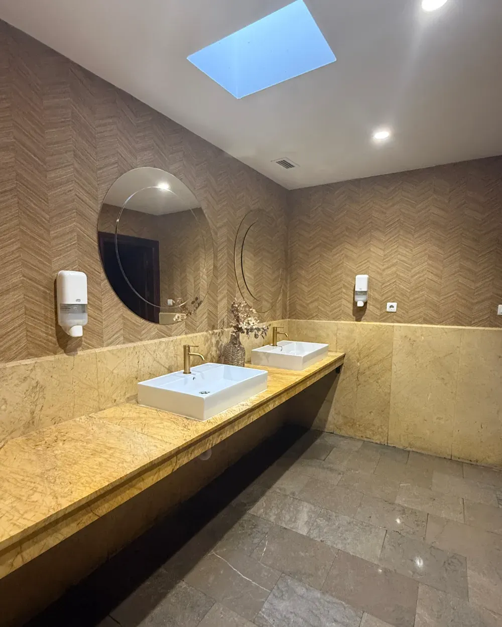 Baño con dos lavabos, encimeras doradas, espejo grande y claraboya. Tonos beige y marrón.