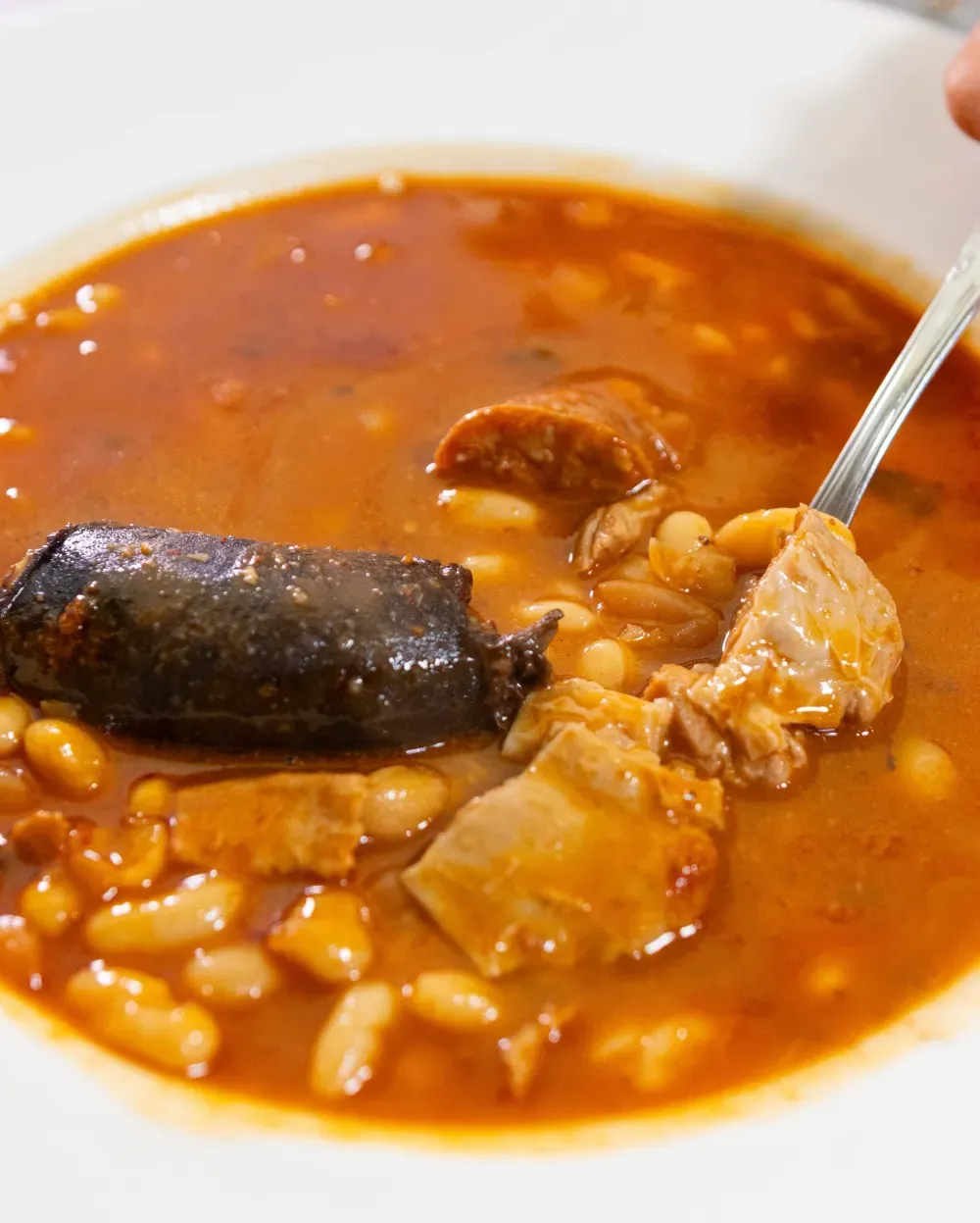 Guiso abundante con frijoles, salchichas y carne en un tazón blanco, sacando un poco con una cuchara.