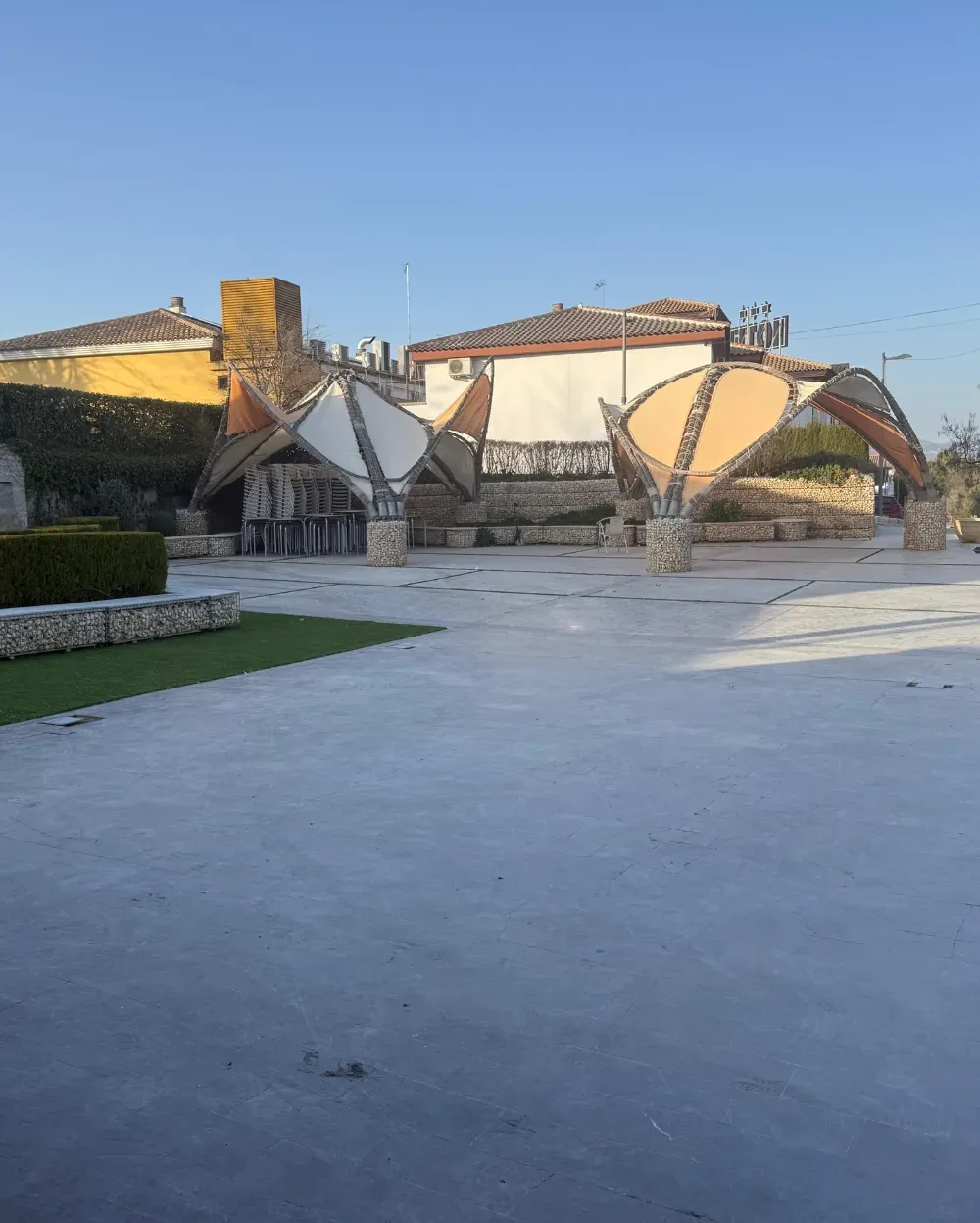 Estructuras de piedra al aire libre con marquesinas arqueadas, sobre una zona pavimentada, con edificios al fondo.
