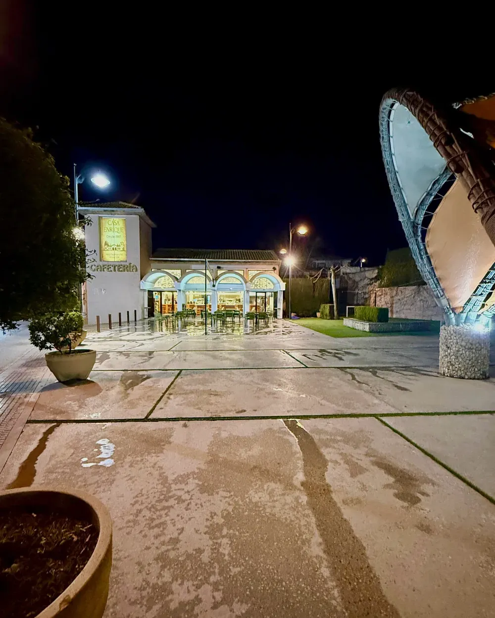 Escena nocturna con un edificio iluminado y una escultura. El pavimento mojado refleja las luces.