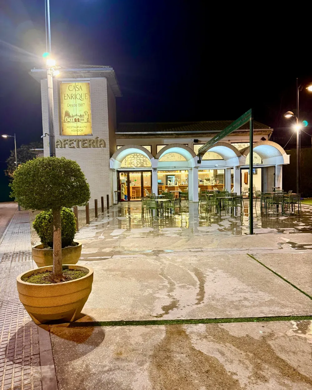 Vista nocturna de un café con asientos al aire libre y ventanas arqueadas bajo brillantes luces de la calle; suelo mojado.