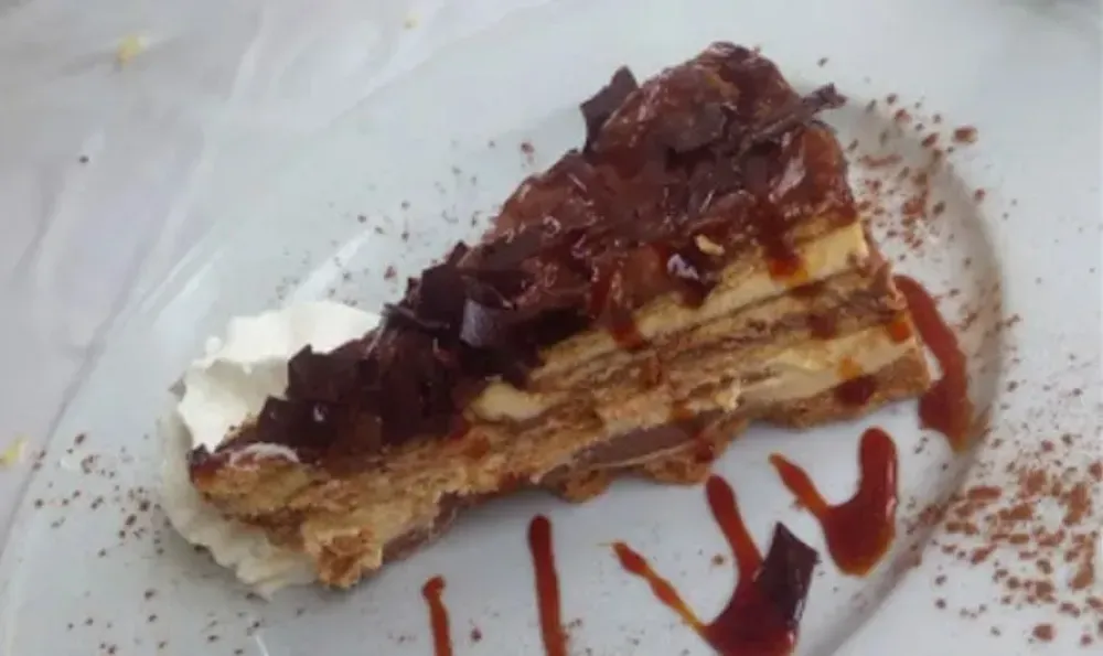 Rebanada de pastel de capas con cobertura de chocolate y caramelo, servida con crema batida en un plato blanco.