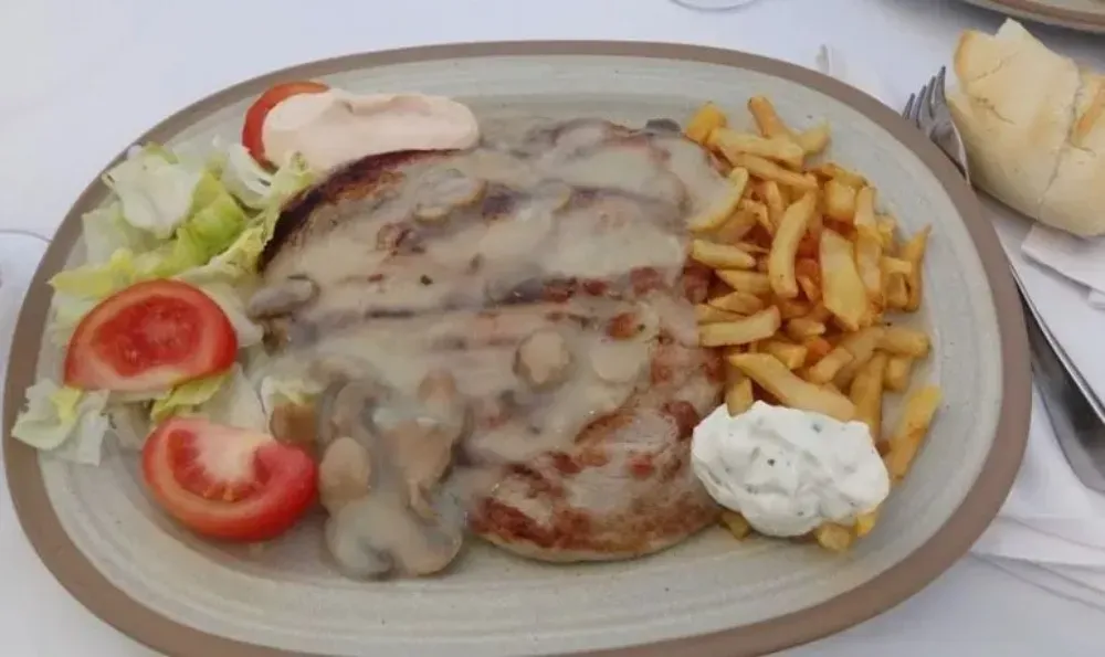 Carne a la parrilla con salsa de champiñones, patatas fritas, tomate, lechuga y salsa para mojar en un plato.