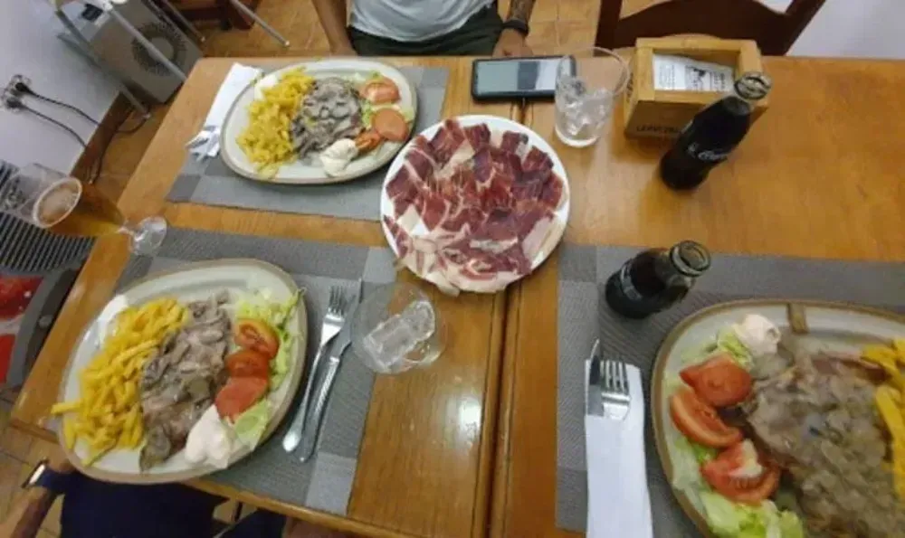 Mesa con platos de comida, jamón, bebidas y cubiertos, preparada para una comida.