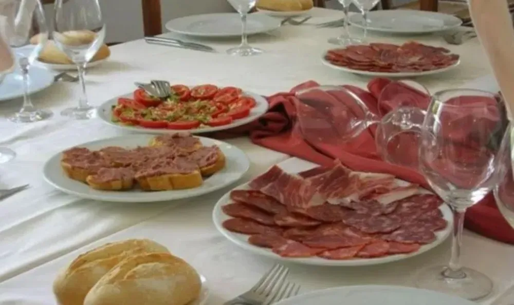 Mesa puesta para una comida: platos de comida que incluyen pan, tomates y carnes cortadas en rodajas, con vasos y mantelería.