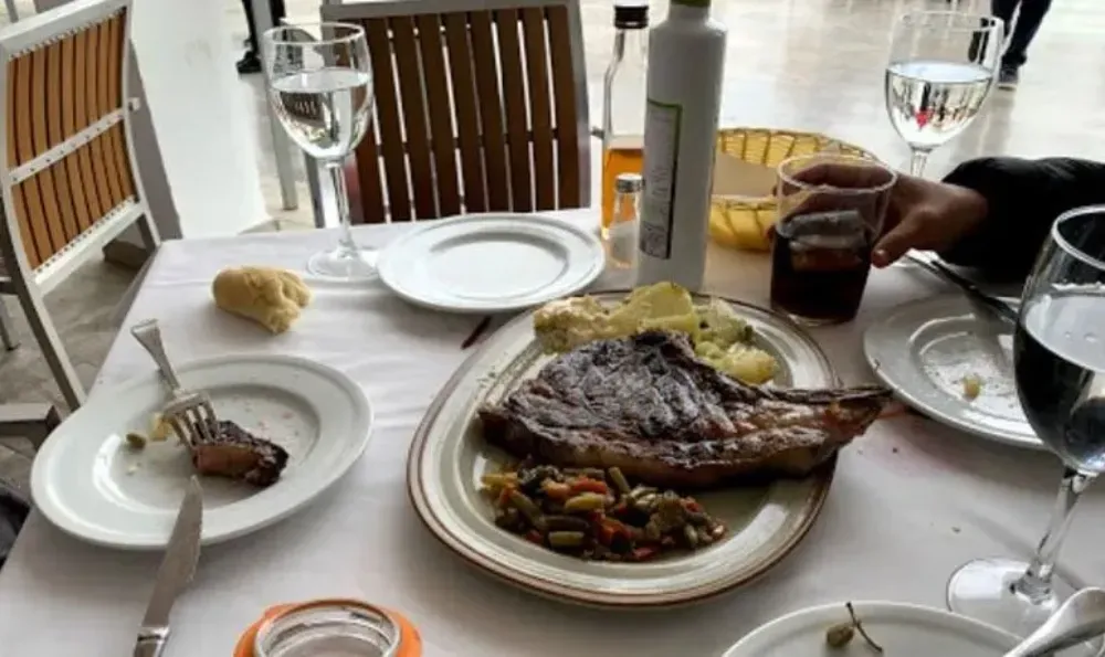 Mesa con filete, verduras, pan, bebidas, platos y vasos. Al aire libre.