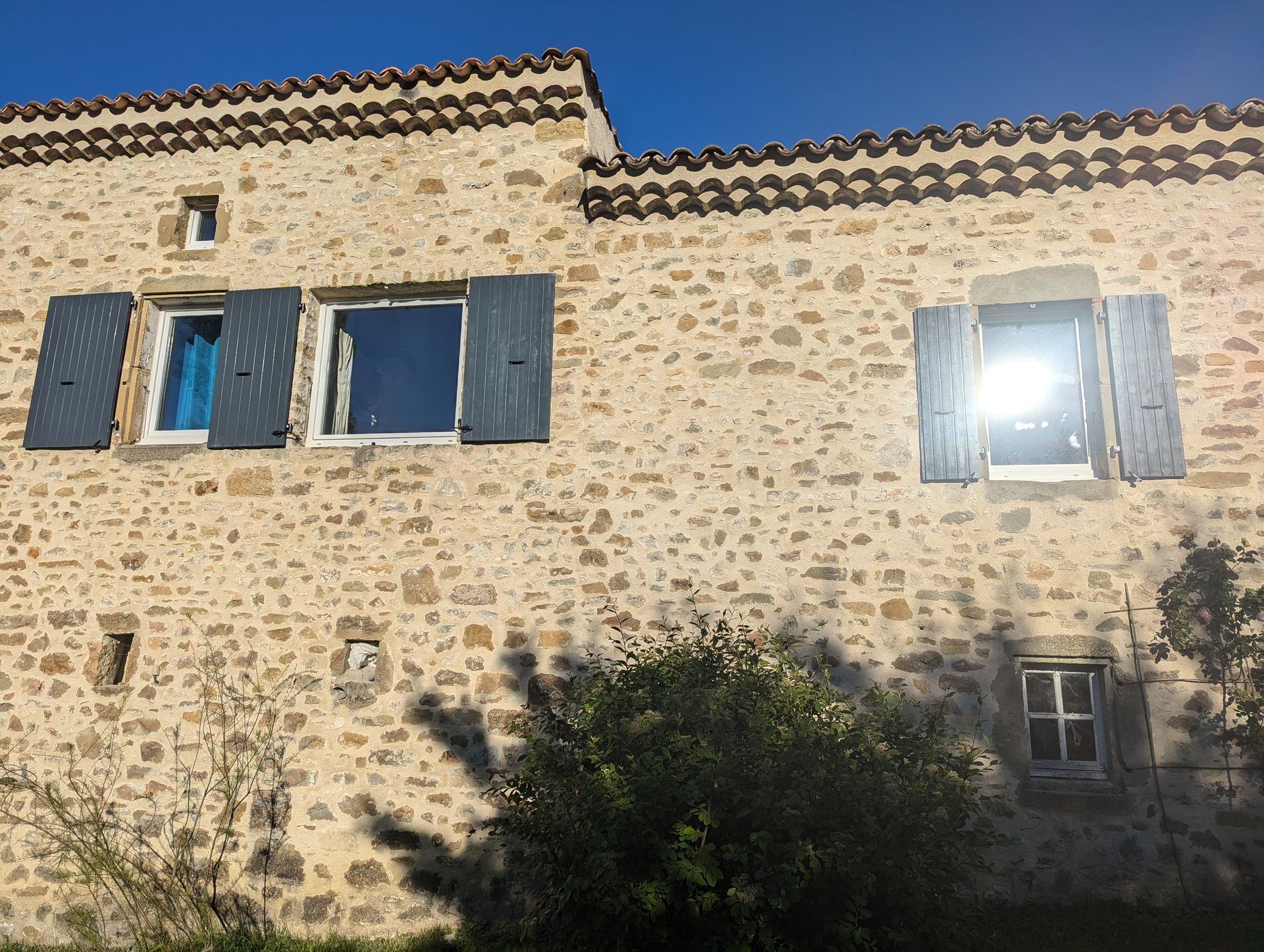 Maison en pierre avec des volets bleus, une petite fenêtre et un reflet ensoleillé dans la vitre.