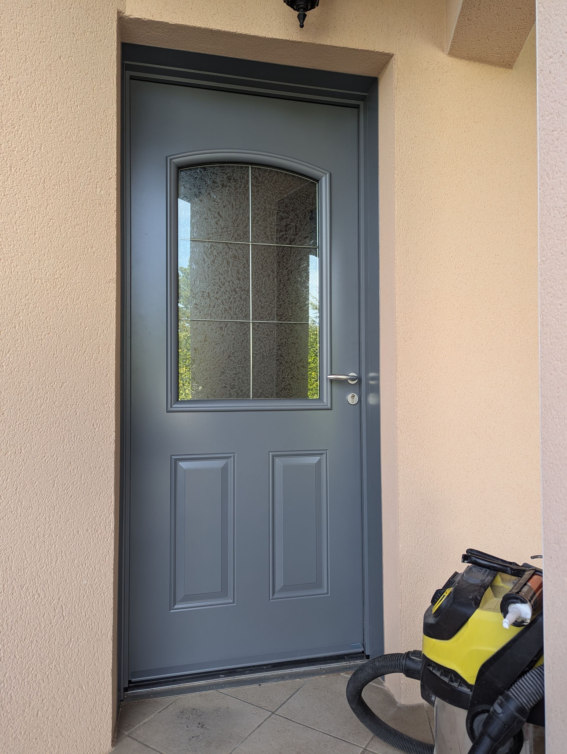 Porte d'entrée grise avec vitrage texturé et panneaux, encastrée dans un mur beige.