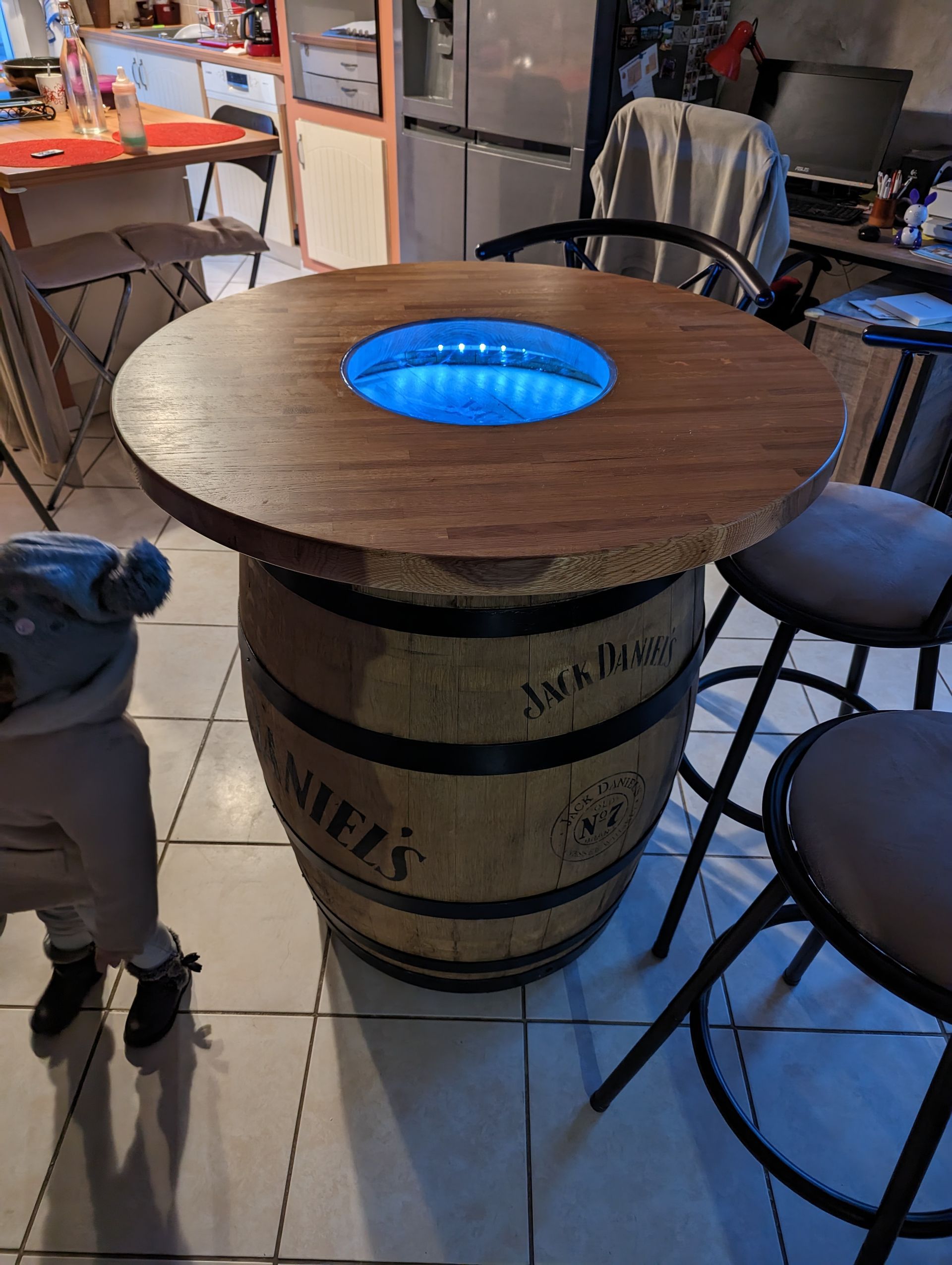 Une table en forme de tonneau avec un effet d'infini bleu lumineux en son centre. Deux tabourets de bar sont disposés à proximité, et un enfant est partiellement visible.