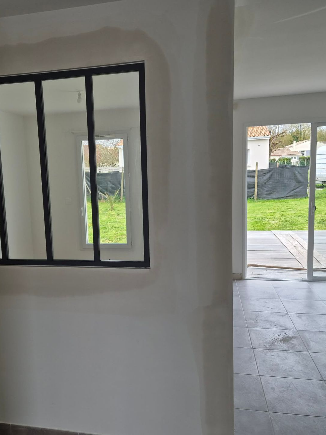 Pièce intérieure avec fenêtre à cadre noir, murs blancs et porte ouverte sur une cour gazonnée.