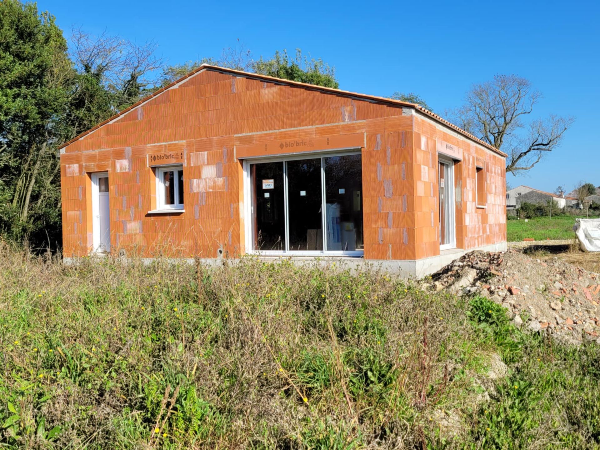 Maison inachevée en briques orange avec portes coulissantes en verre dans un champ herbeux à la campagne, sous un ciel dégagé