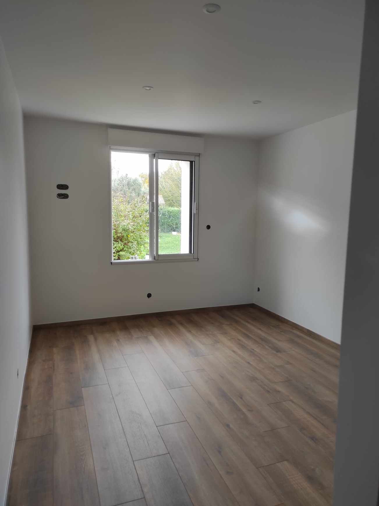 Chambre vide non meublée avec murs blancs, parquet et une fenêtre laissant entrer la lumière du jour