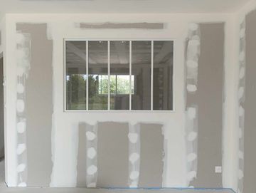 Mur intérieur inachevé avec des plaques de plâtre réparées et une fenêtre laissant entrer la lumière du jour