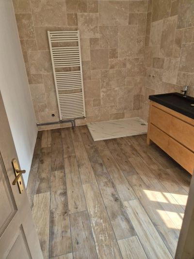 Salle de bains vide avec sol imitation bois, mur carrelé en pierre, radiateur blanc et meuble-lavabo en bois.
