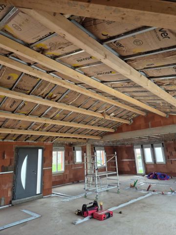 Intérieur d'une maison en briques en construction, avec poutres de toit apparentes, échafaudages et outils au sol.
