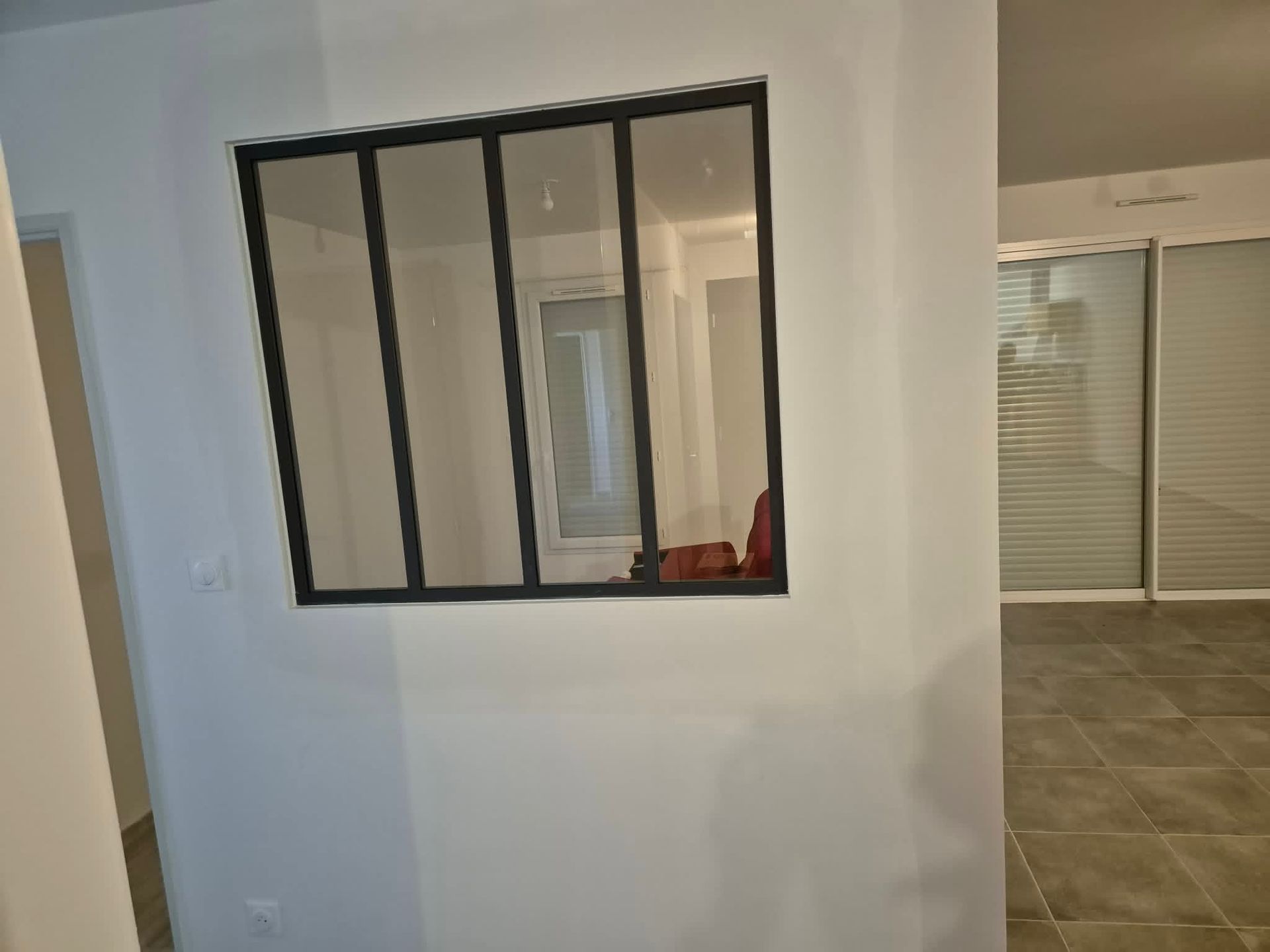 Pièce vide aux murs blancs, fenêtre à cadre noir et portes coulissantes en verre donnant sur une cour gazonnée.