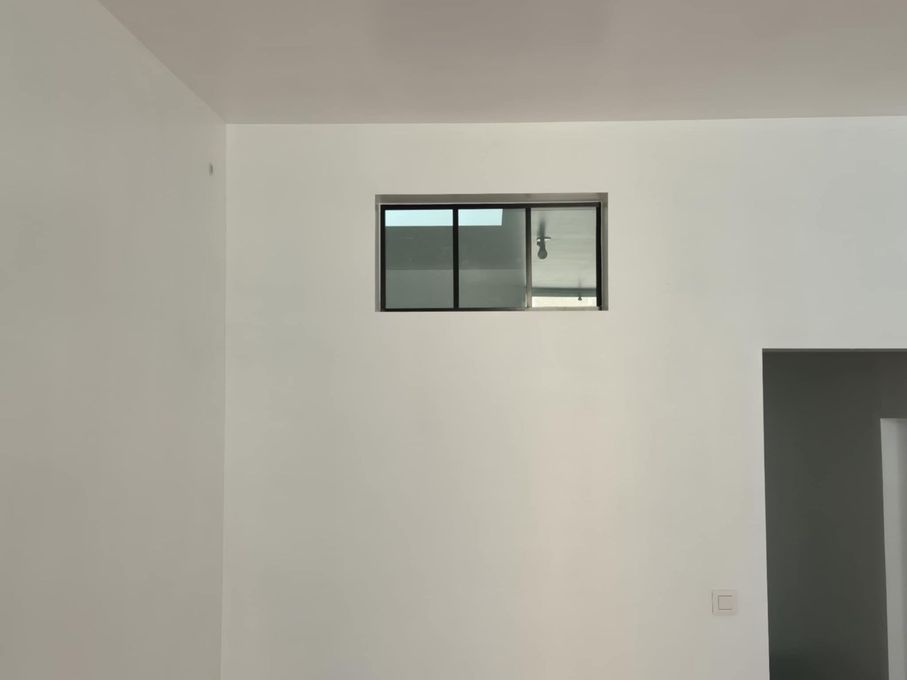 Pièce blanche vide avec une petite fenêtre rectangulaire en hauteur sur le mur et une porte ouverte sur la droite.