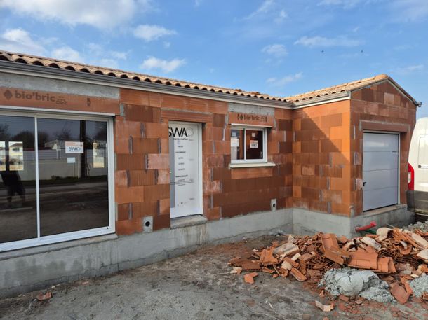 Maison de plain-pied en construction, avec murs en briques, porte blanche, grande fenêtre et tas de débris à l'extérieur.