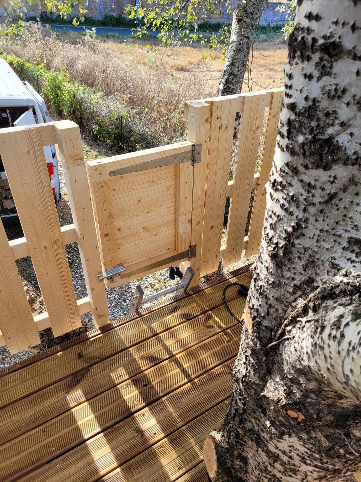 Portail en bois et terrasse à côté d'un arbre, donnant sur une cour ensoleillée.