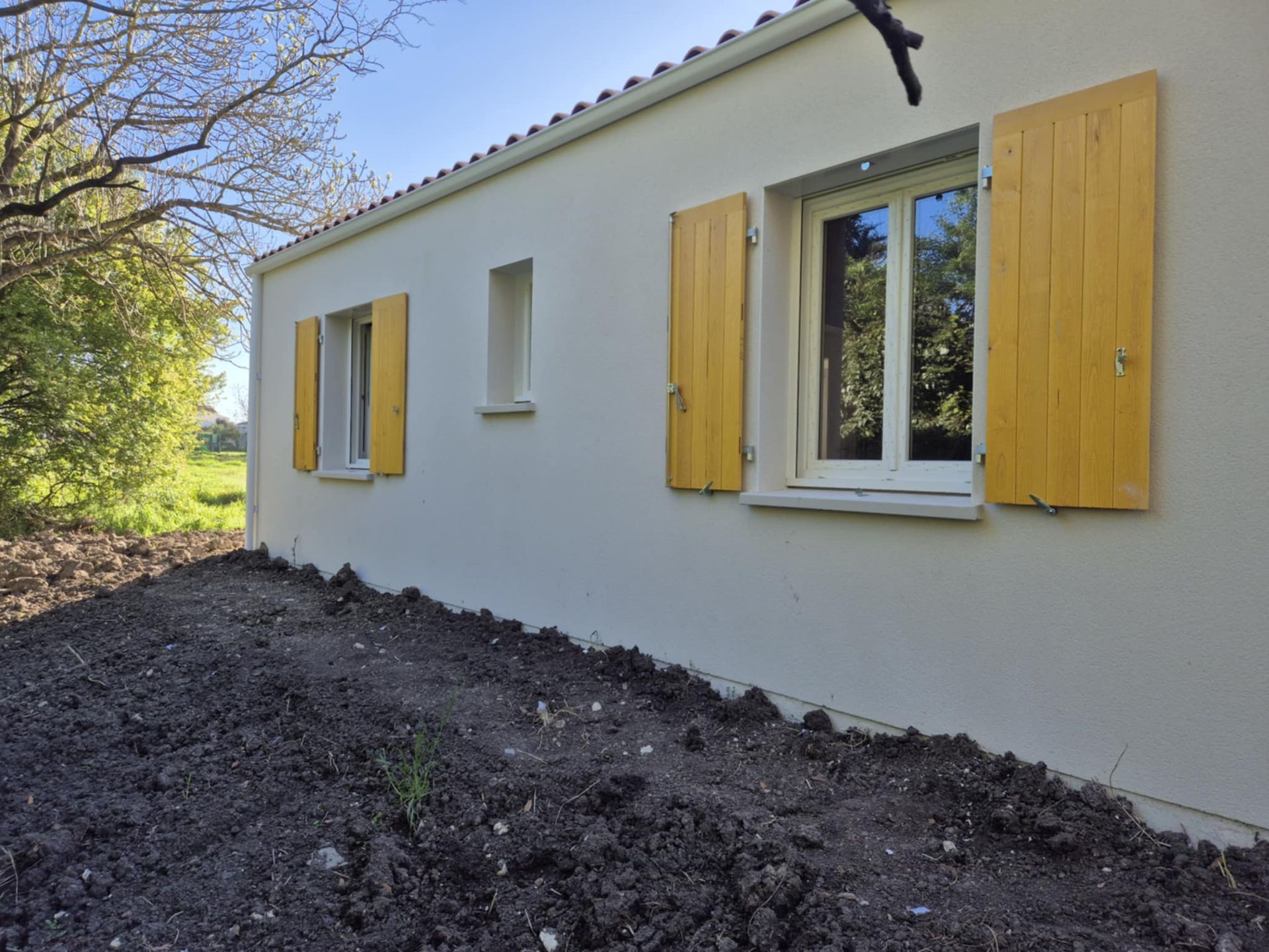 Maison blanche avec volets jaunes, à côté d'une cour boueuse et d'arbres.