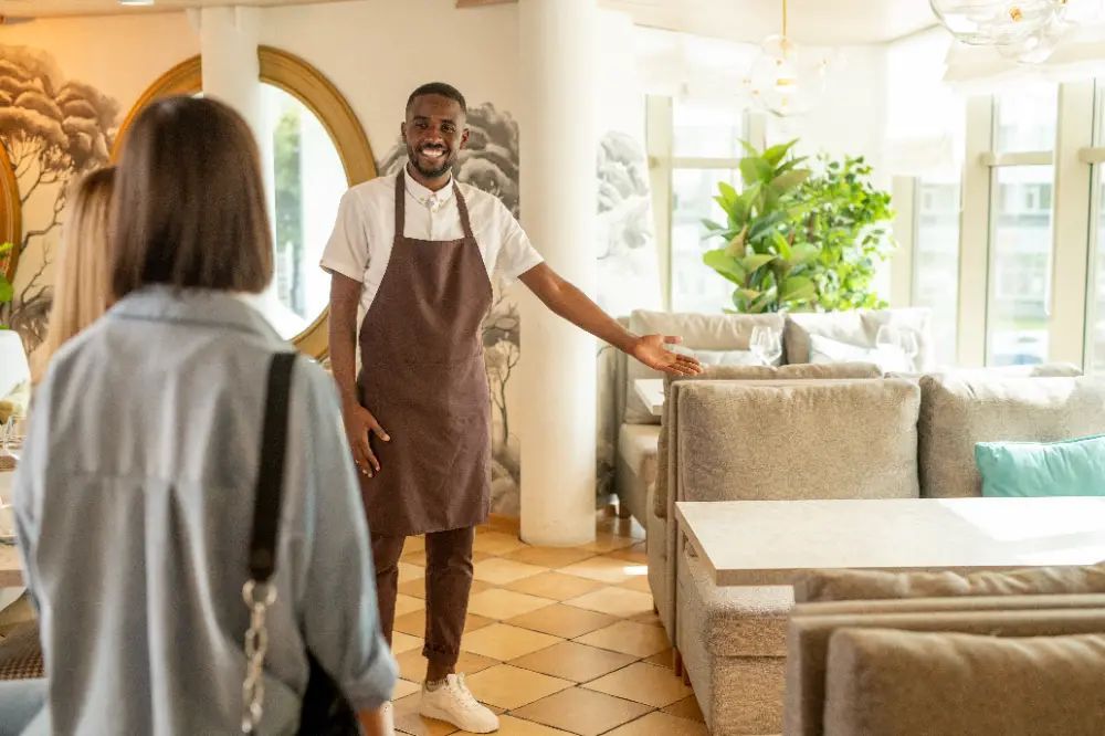 A man in an apron is standing in a restaurant talking to a woman.