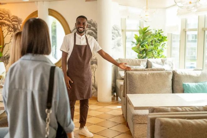 A man in an apron is standing in a restaurant talking to a woman.