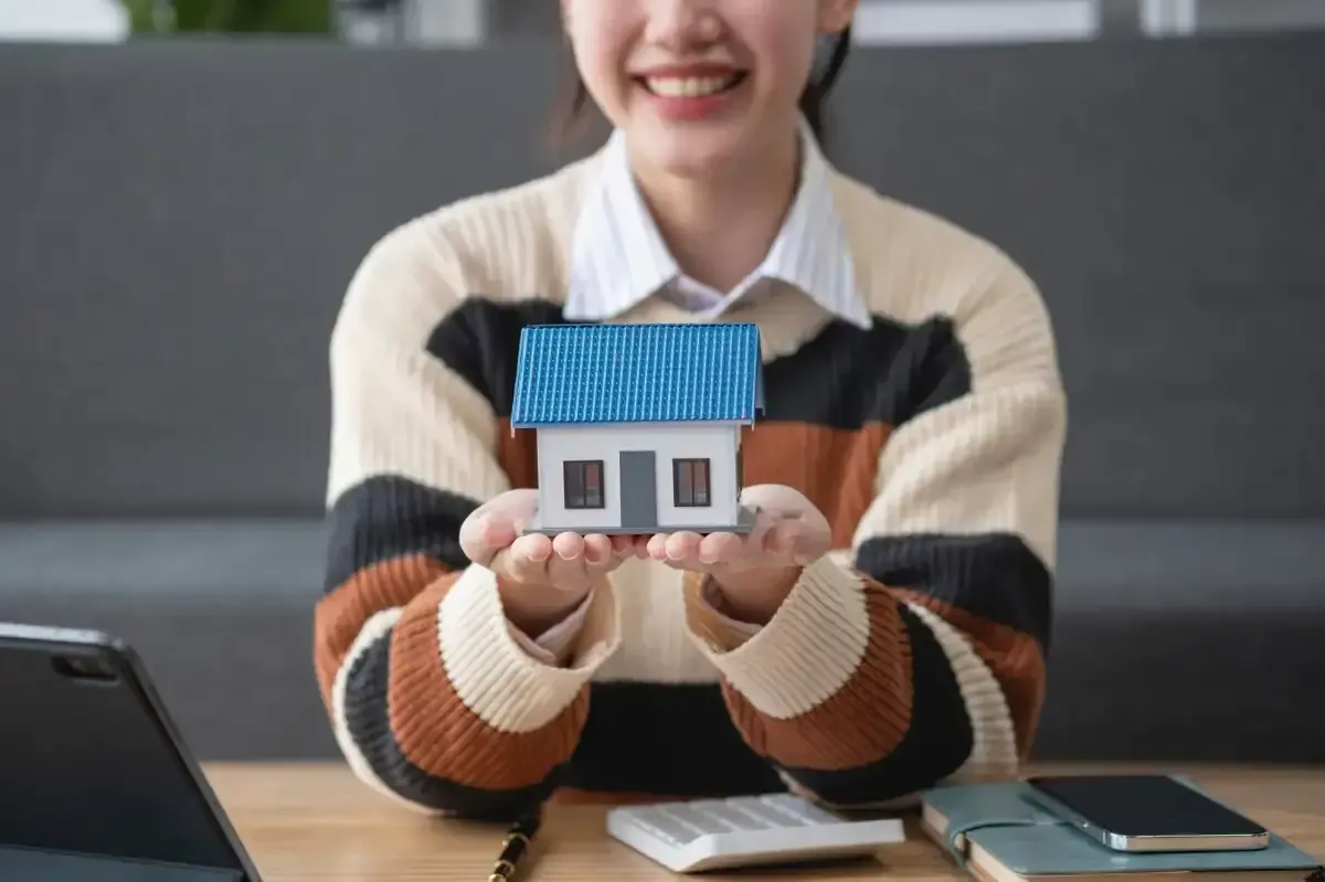 Una persona sonriente sostiene una casa en miniatura, que posiblemente simboliza la propiedad de una vivienda o de bienes raíces.