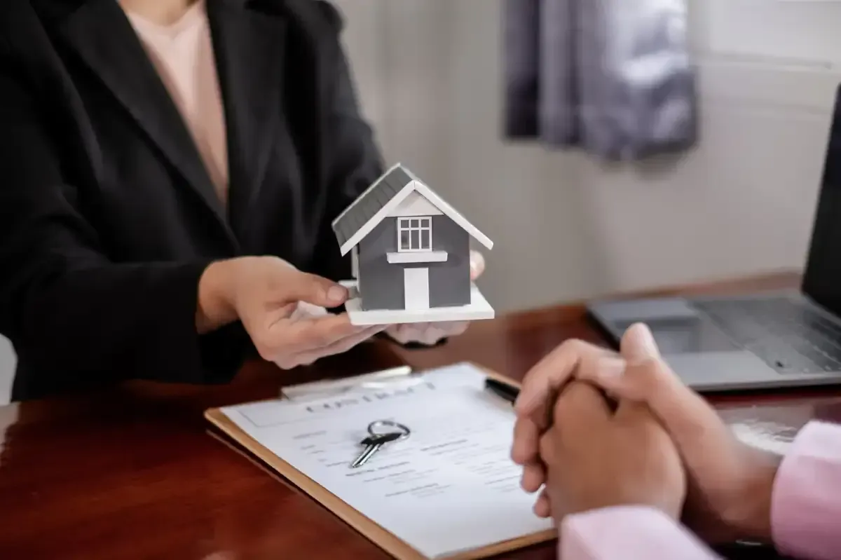 Persona sosteniendo una maqueta de casa sobre papeleo, con las manos entrelazadas.