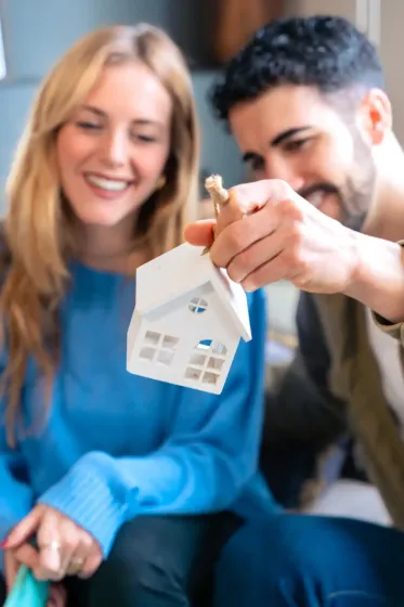Pareja sonriendo, mirando una pequeña maqueta de casa sostenida por el hombre, probablemente una casa nueva.