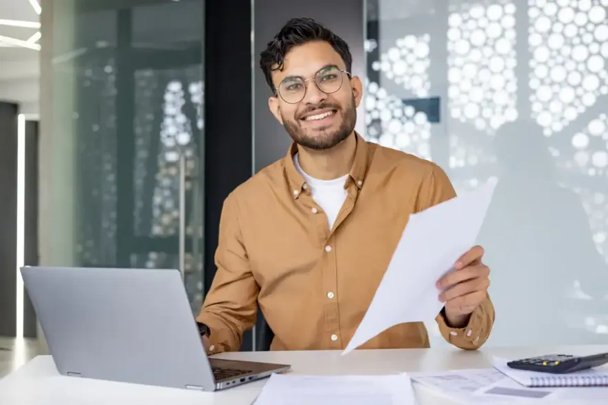 Un hombre con gafas sonríe, sostiene papeles y usa una computadora portátil en el escritorio de la oficina.