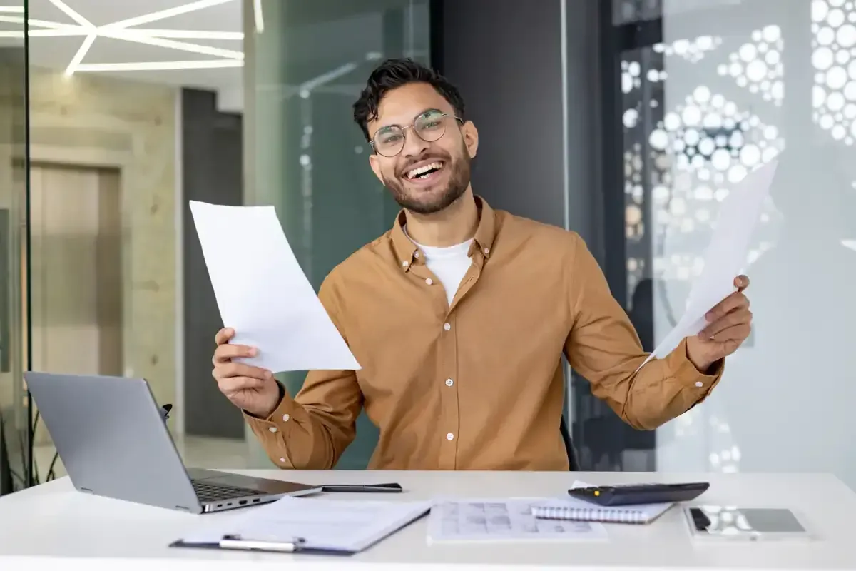 Hombre con camisa marrón, sonriendo, sosteniendo papeles en un escritorio con computadora portátil, calculadora y cuaderno.