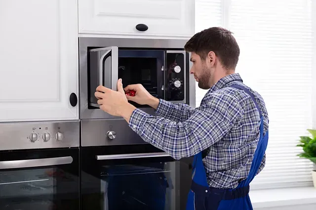 Hombre con ropa de trabajo reparando un horno microondas en una cocina blanca.