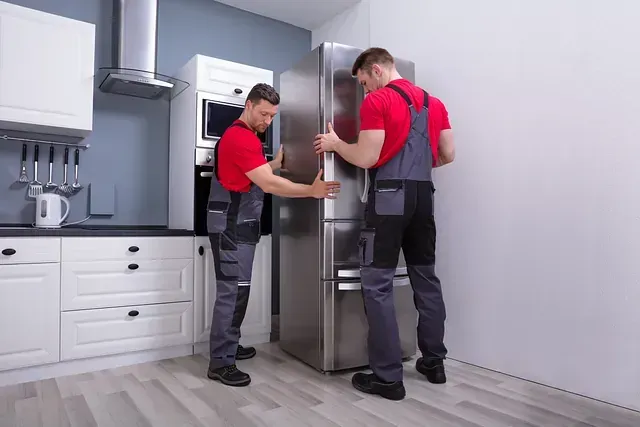 Dos operarios con camisas rojas y monos grises instalando un refrigerador de acero inoxidable en una cocina moderna.