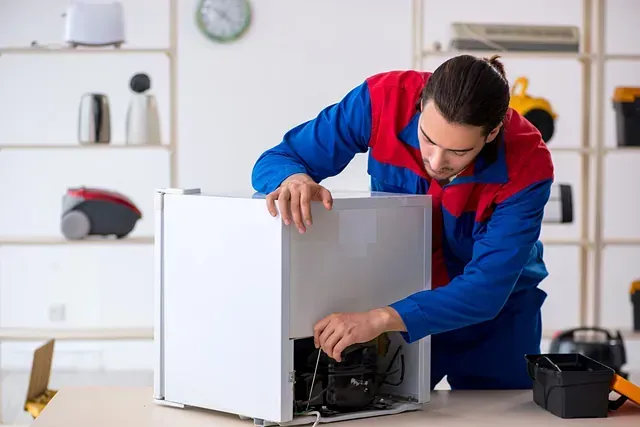 Un hombre con uniforme de trabajo repara un mini-refrigerador blanco sobre una mesa en un taller bien iluminado.