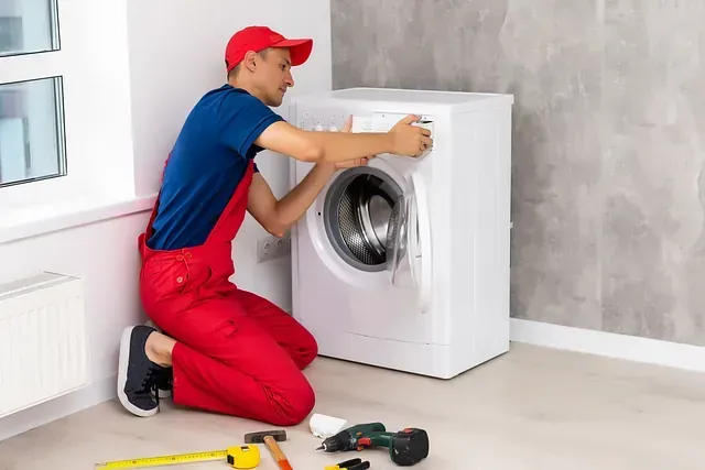 Un hombre con un mono rojo y una gorra repara una lavadora blanca en el interior de una casa.