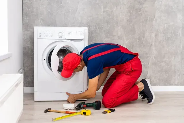 Hombre con mono rojo reparando una lavadora, arrodillado en el suelo con herramientas.