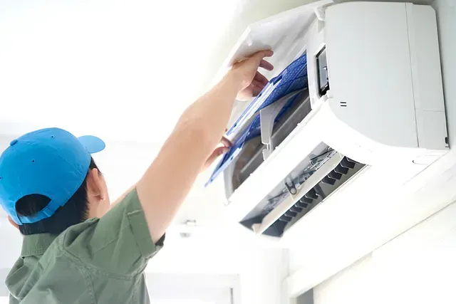 Una persona con una gorra azul limpiando el filtro de aire de una unidad de aire acondicionado blanca montada en una pared blanca.