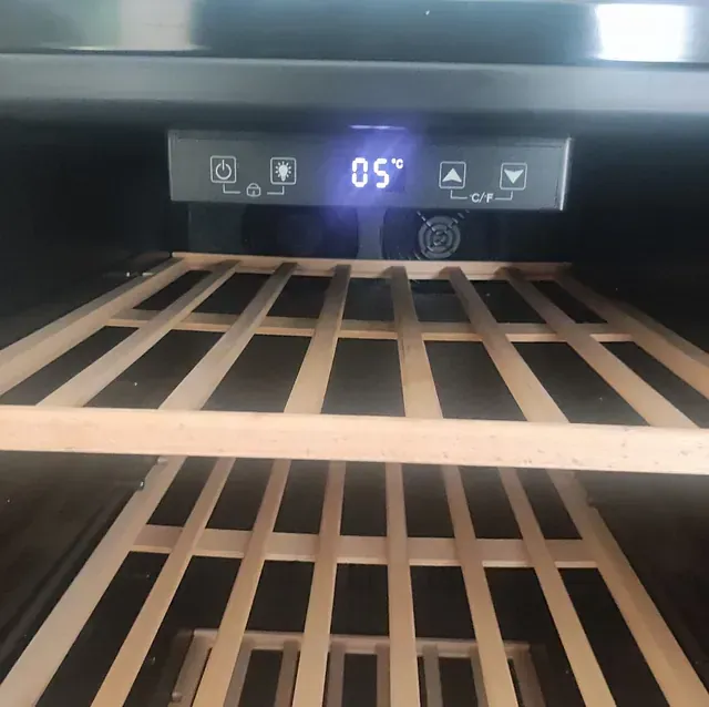 Dentro de un enfriador de vinos, estantes de madera vacíos y una pantalla digital que muestra 05°C.