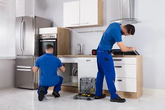 Dos hombres con uniformes azules instalan gabinetes de cocina, uno arrodillado y el otro de pie.