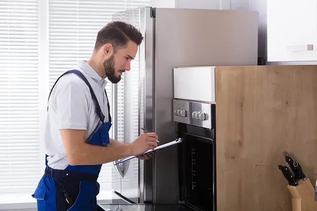 Un técnico con mono azul inspecciona el horno, tomando notas en una cocina con electrodomésticos de acero inoxidable y una ventana.