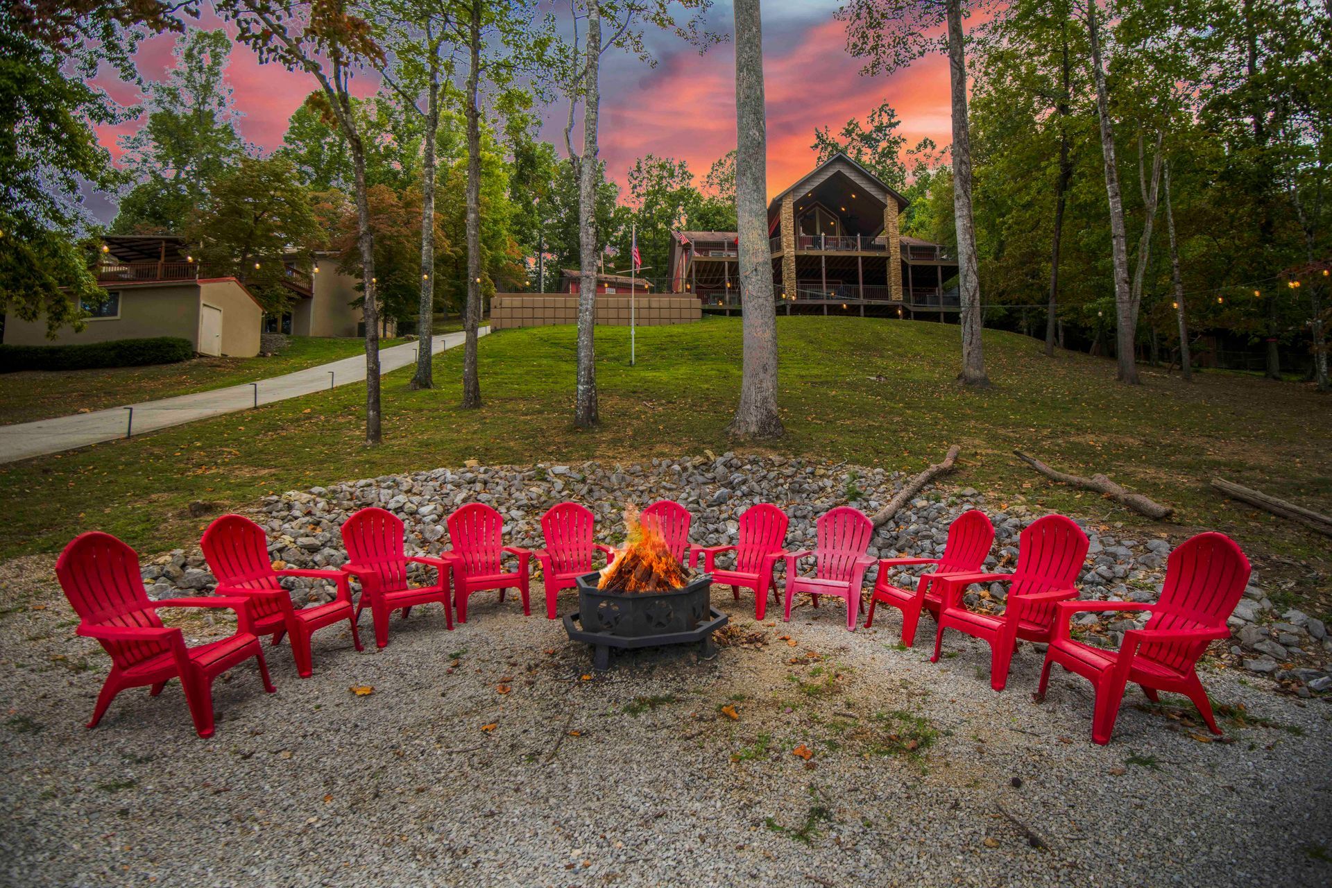 Red chairs circle a fire pit on a gravel area, with a house and trees in the background at sunset.