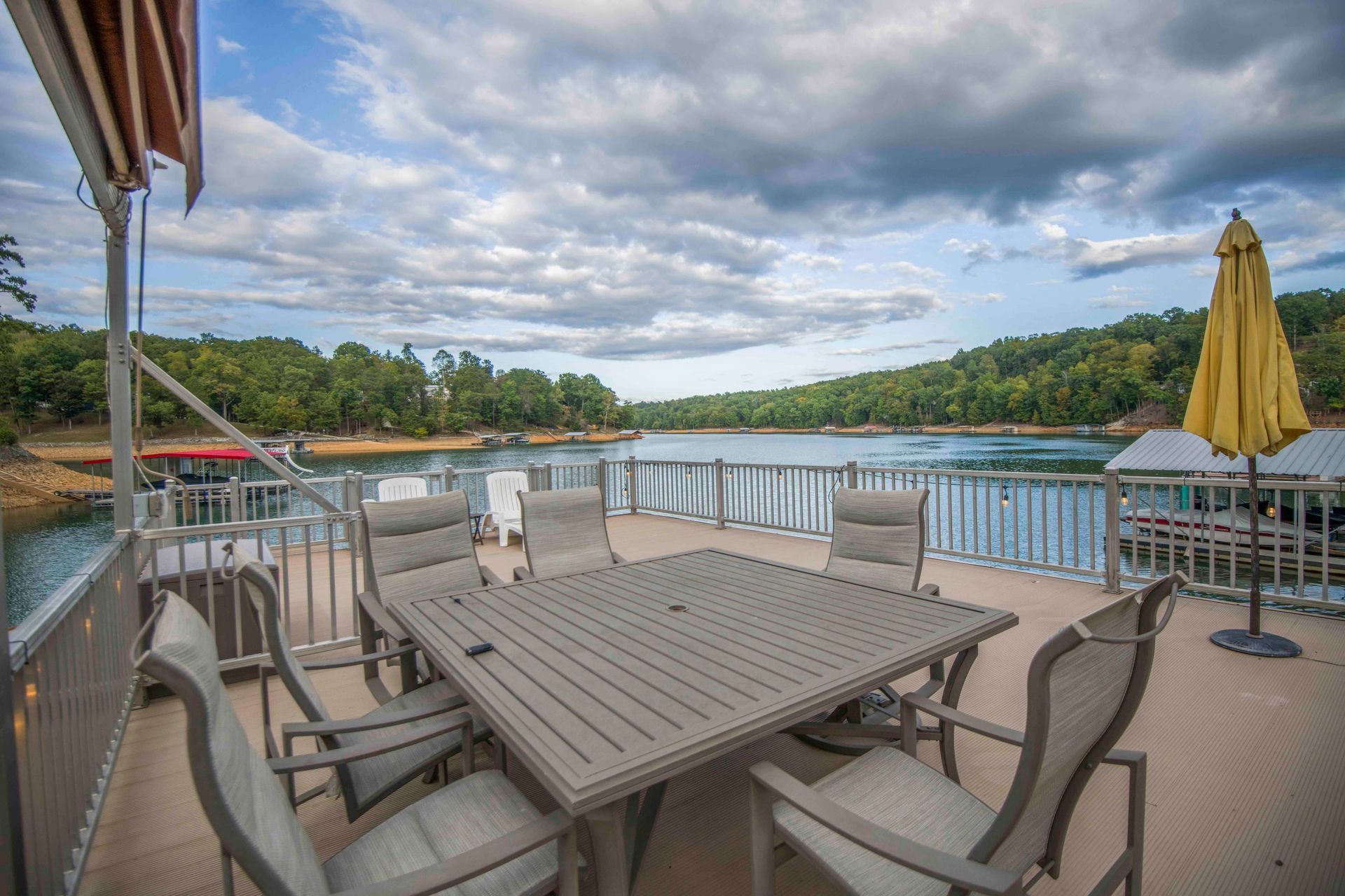 Outdoor dining set on a houseboat deck overlooking a lake surrounded by trees. Cloudy sky.