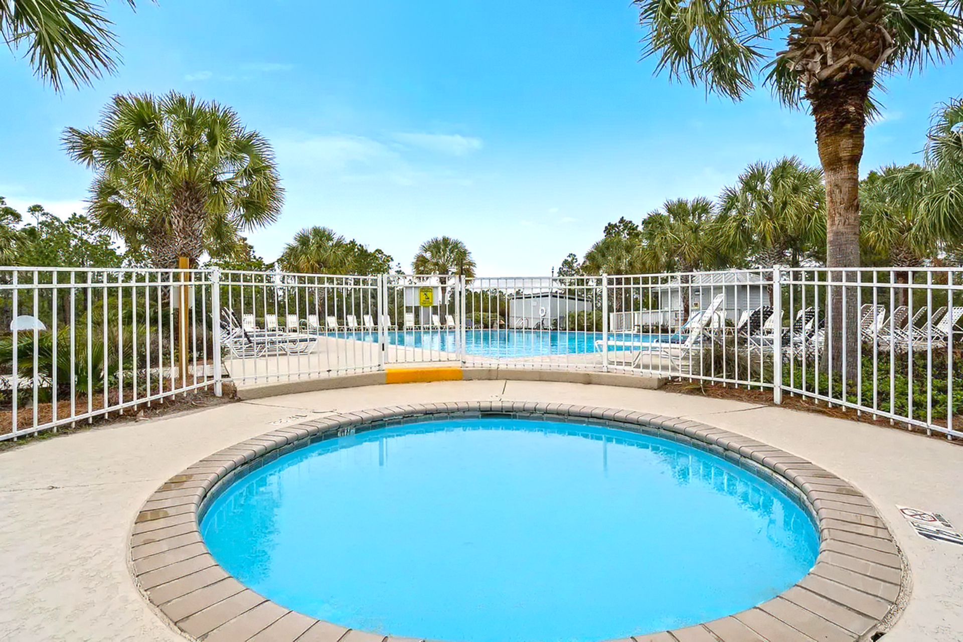 Small round pool with blue water, white fence, and palm trees in a sunny courtyard