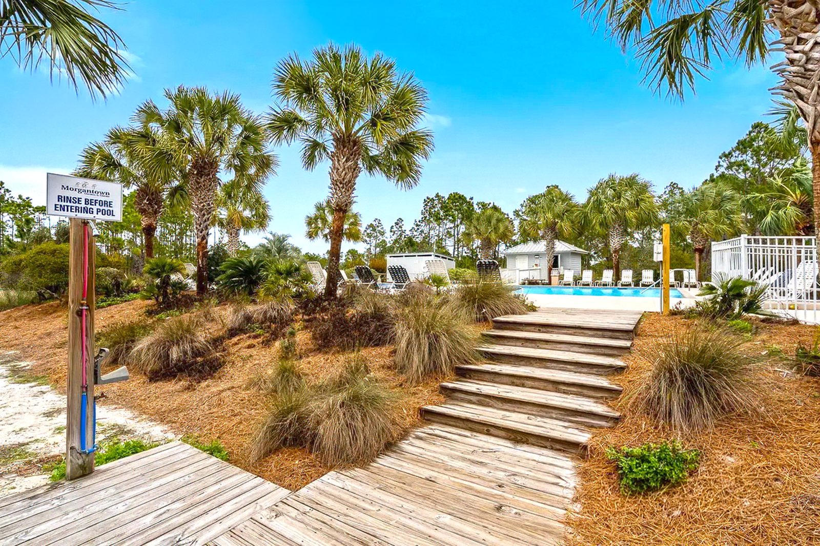 Wooden boardwalk steps through dunes with palm trees and a pool in the background
