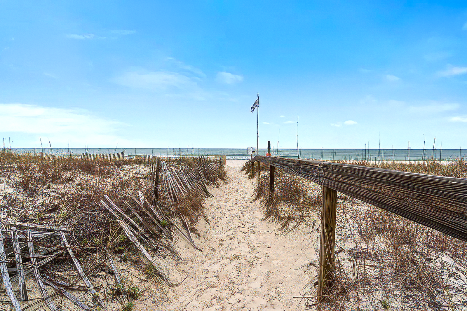 Sandy beach path between dune fences leading to the ocean under a clear sky
