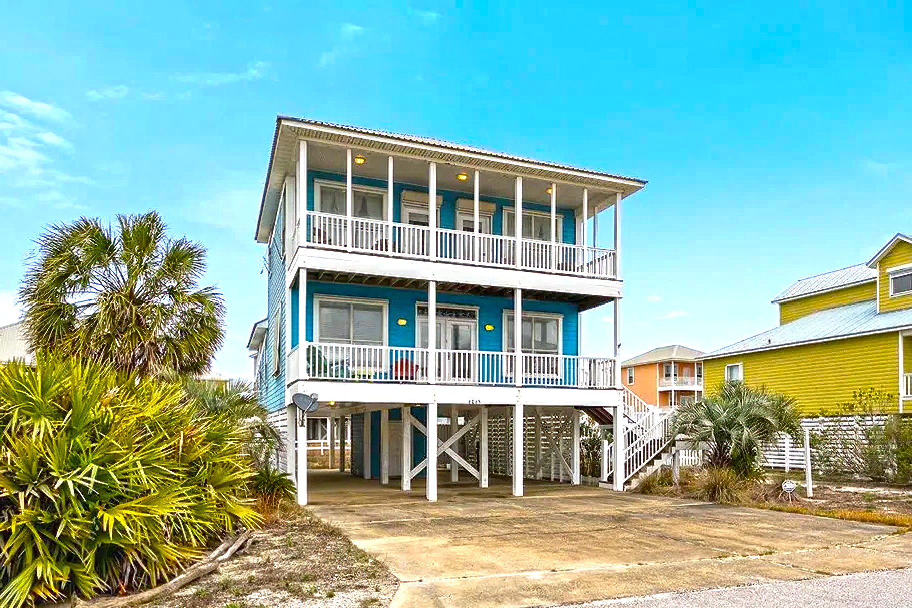 Elevated beach house with white railings and blue shutters beside sandy dunes under a bright blue sky