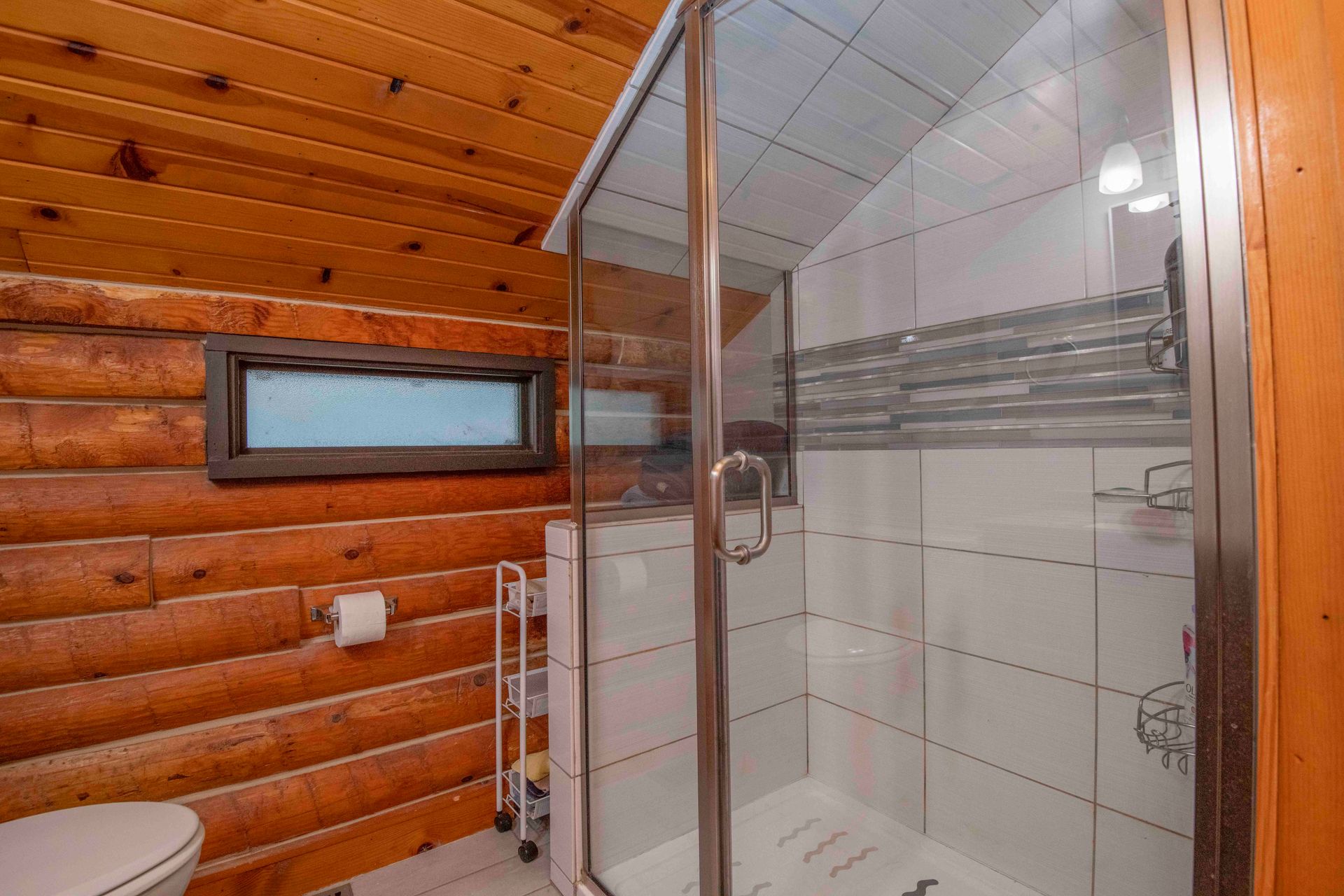 Bathroom with a glass shower door, white tile, wooden walls, and a small window.