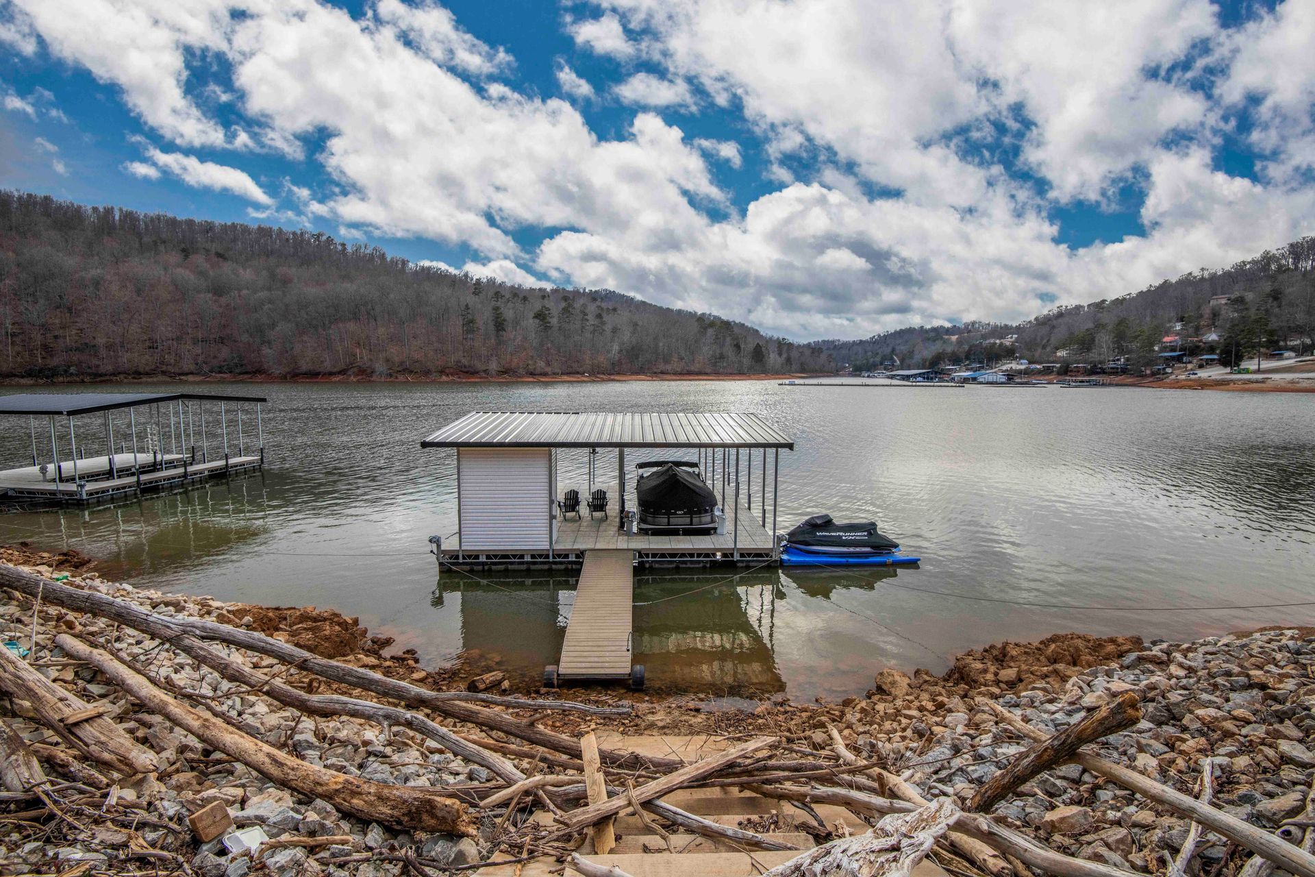 Lake scene with a boat dock and a boat, under a blue and cloudy sky. Hills surround the lake.