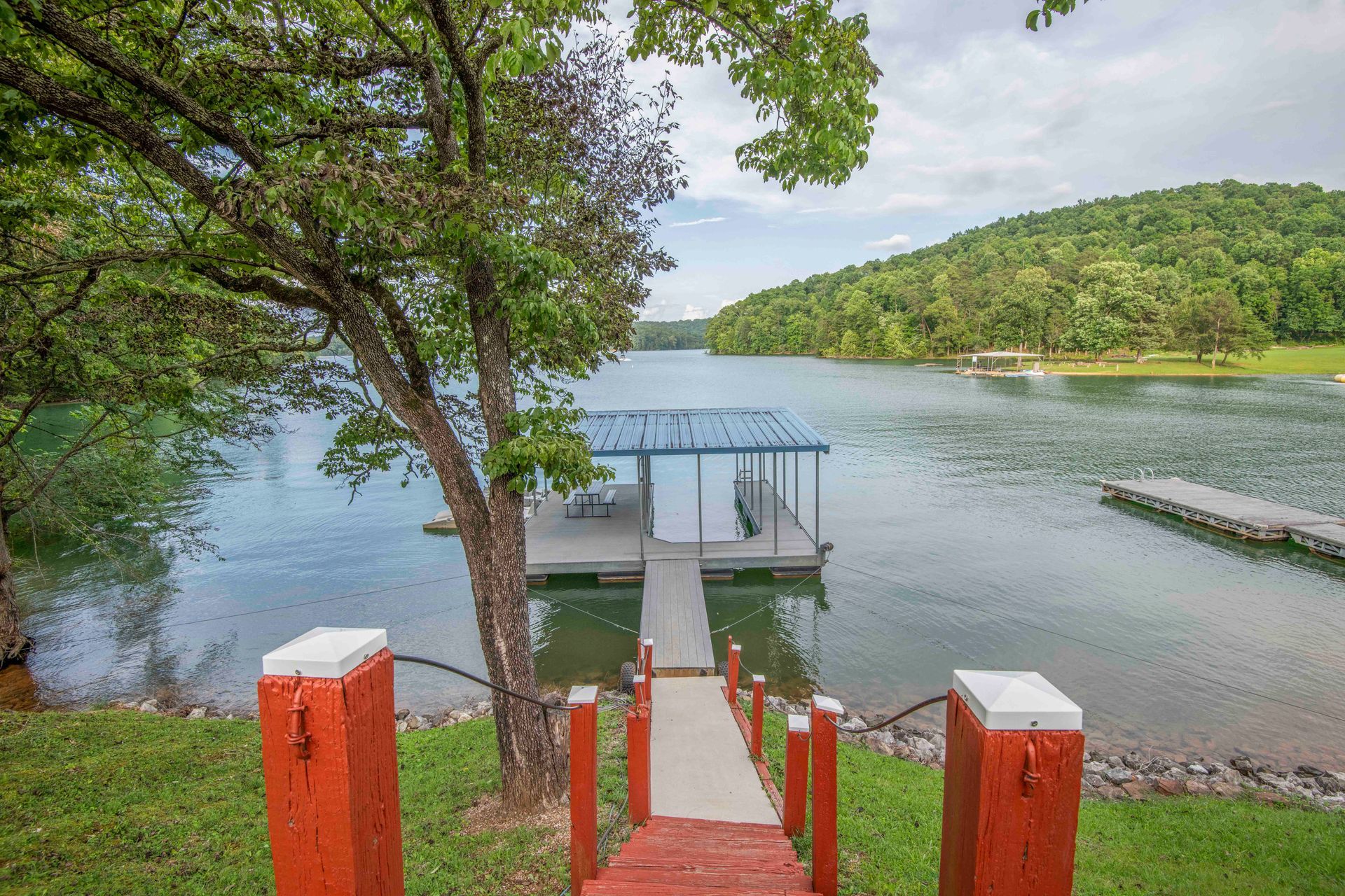 Wooden dock with a boathouse on a lake, viewed from stairs descending from a grassy yard.